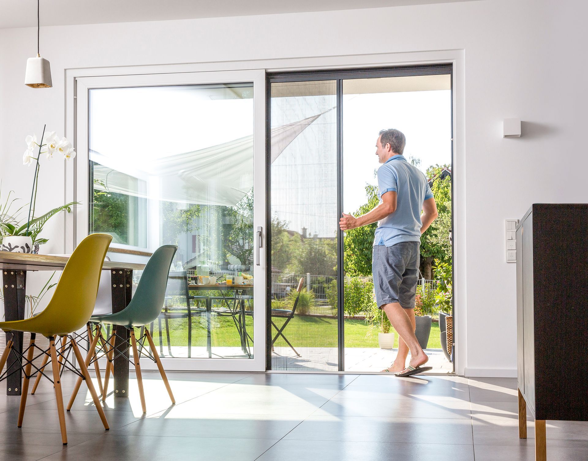 Mann öffnet eine Glasschiebetür mit einziehbarem Fliegengitter, sonniger Innenraum mit Tisch und Stühlen.