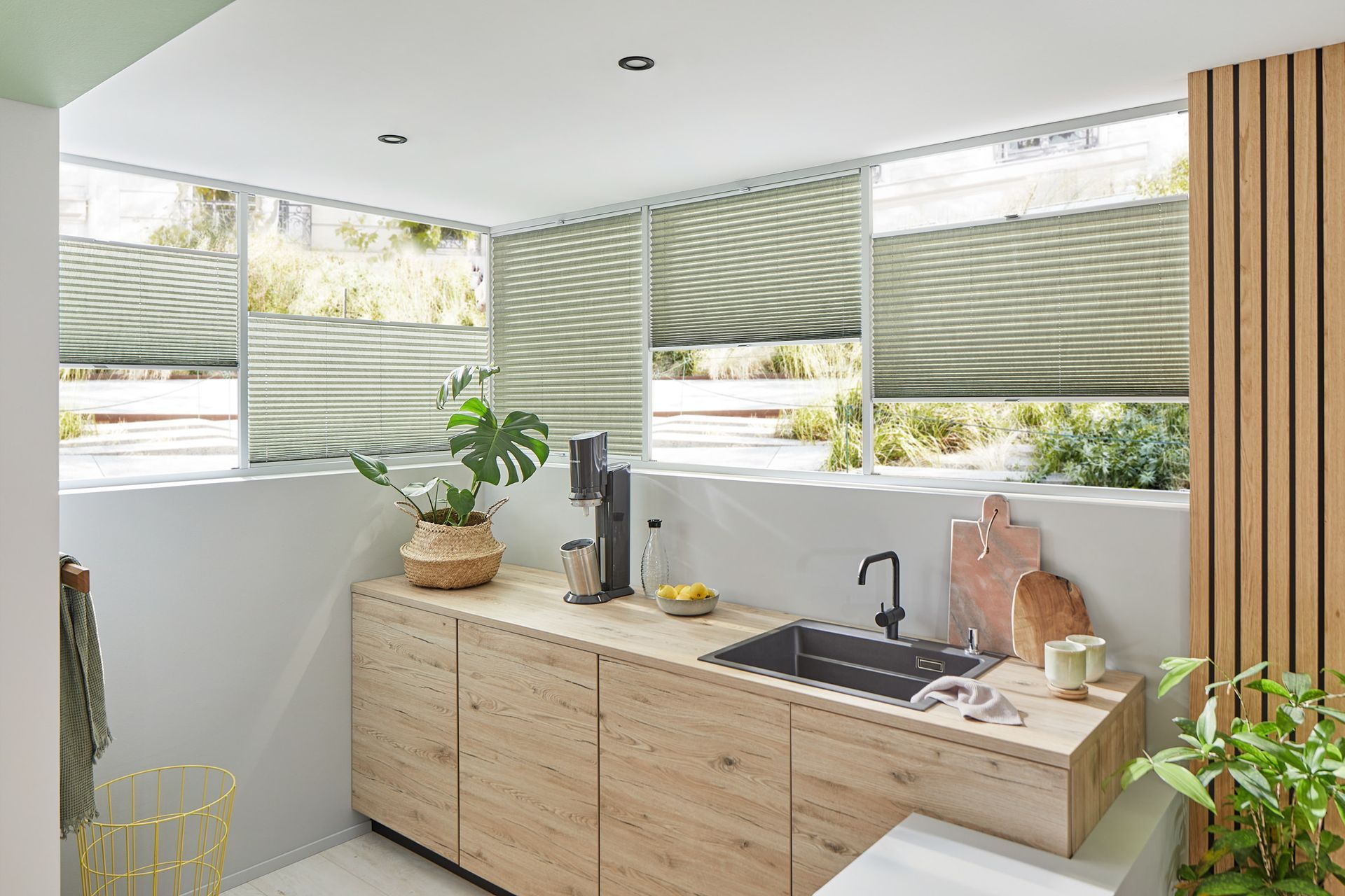 Küche mit hellen Holzschränken, schwarzem Spülbecken und hellgrünen Jalousien an den Fenstern.