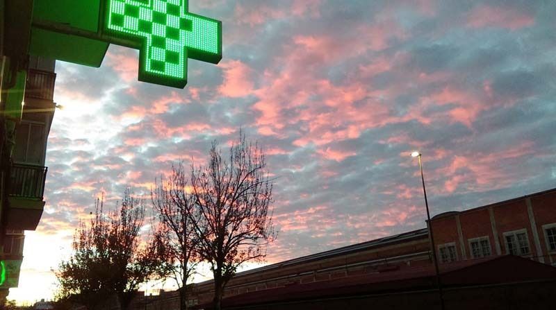 Una cruz verde cuelga del costado de un edificio con una puesta de sol de fondo.