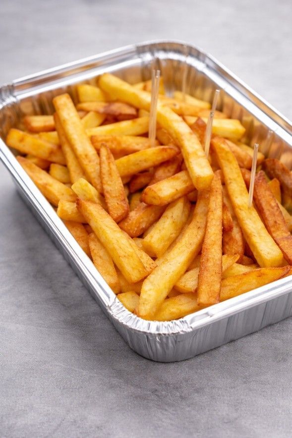 Una ración de patatas fritas doradas en un envase rectangular de aluminio para llevar, sobre una superficie gris.