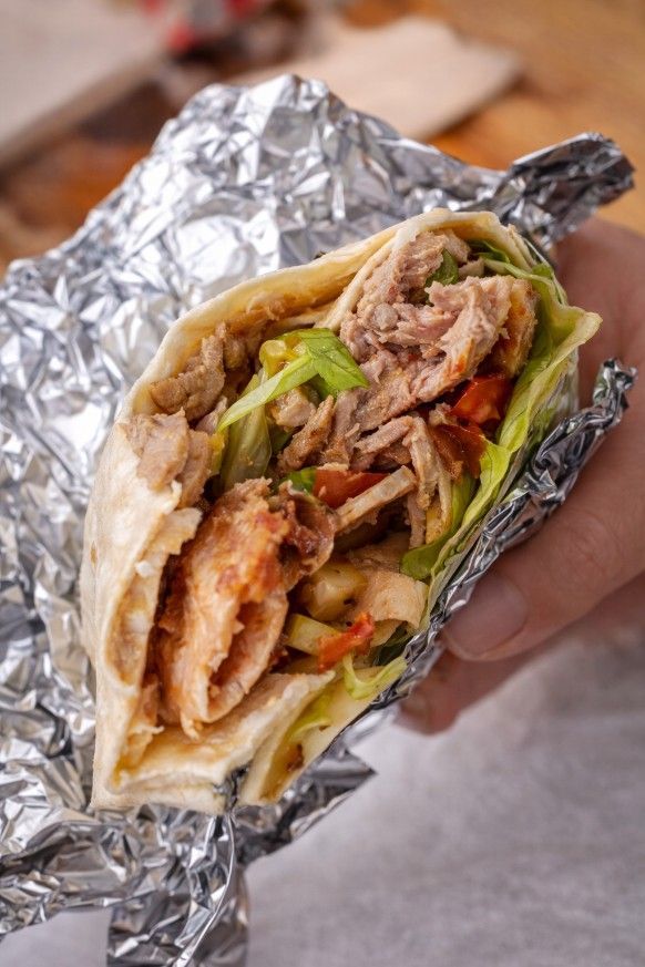Una mano sostiene un wrap de carne y verduras parcialmente desenvuelto en papel de aluminio arrugado.