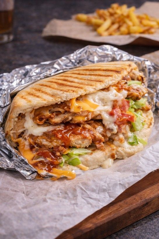 Un sándwich de pollo prensado envuelto en papel de aluminio, servido sobre papel pergamino con una guarnición de papas fritas