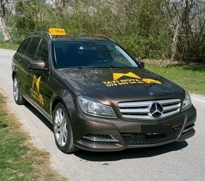 Ein braunes Mercedes-Benz Taxi mit der Aufschrift „TAXI NOVA“ und einer Telefonnummer auf der Motorhaube parkt auf einem Parkplatz im Freien.