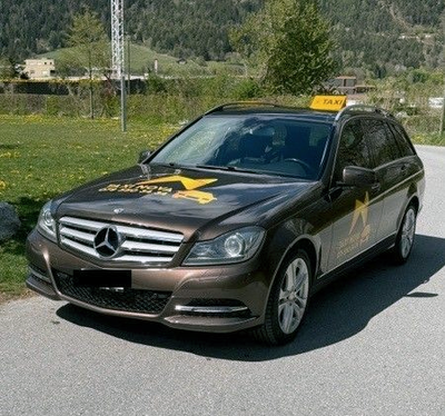 Ein dunkelbraunes Mercedes-Benz-Taxi parkte tagsüber auf einem Parkplatz; im Hintergrund war ein mehrstöckiges Wohnhaus zu sehen.