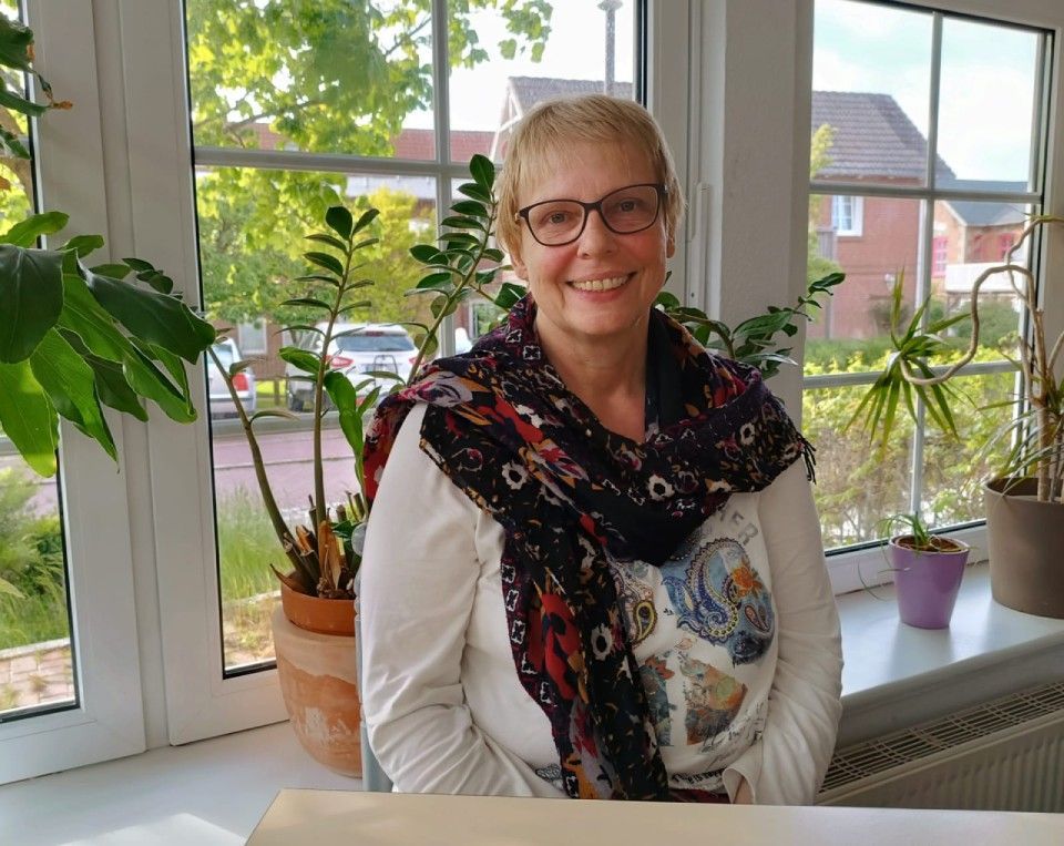 Eine Frau mit Brille, Schal und weißem Hemd lächelt in der Nähe eines Fensters mit Pflanzen.