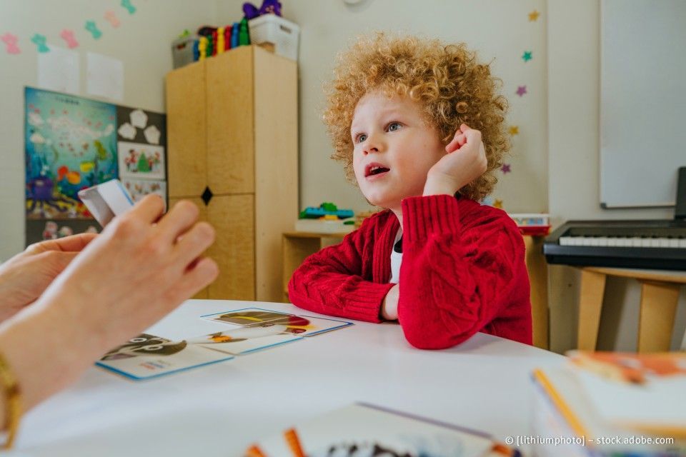Kind mit lockigem Haar schaut während des Unterrichts in ein Buch, roter Pullover, drinnen.