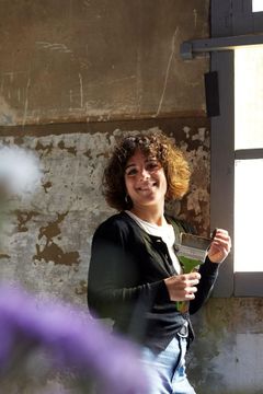 Persona amb els cabells arrissats somrient i sostenint una beguda a l'interior al costat d'una finestra assolellada