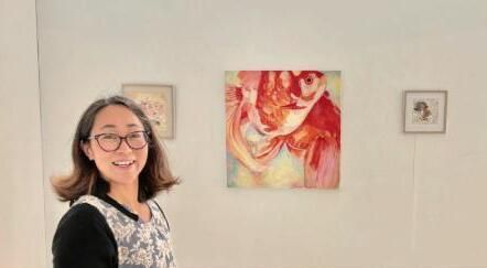 Una dona somrient amb ulleres està al costat de tres quadres abstractes emmarcats en una paret color crema.