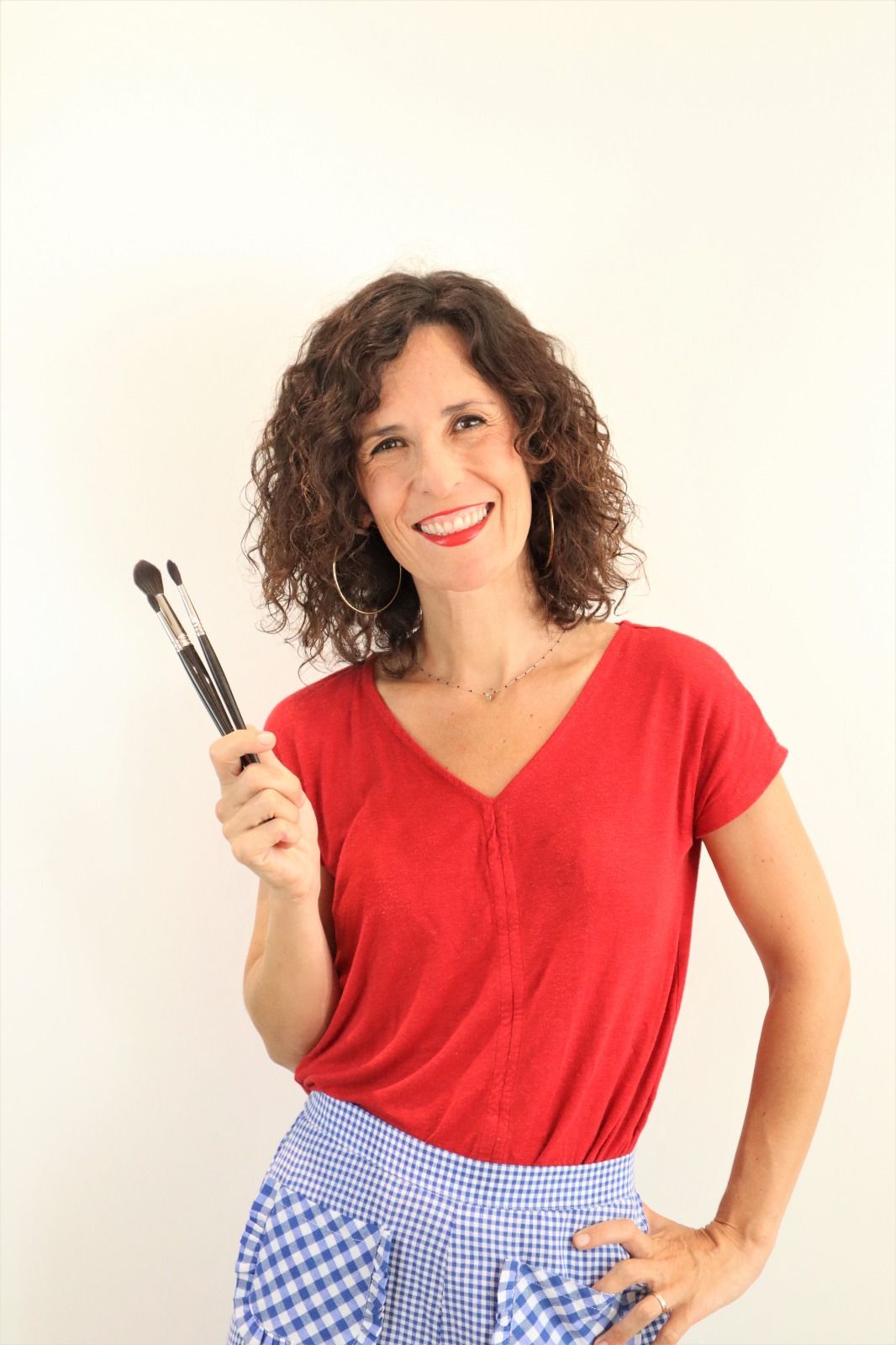 Dona que sosté pinzells de maquillatge, somrient, amb camisa vermella i faldilla blava de quadres vichy.