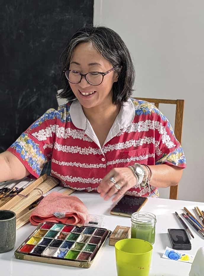 Una dona somrient amb ulleres està al costat de tres quadres abstractes emmarcats en una paret color crema.
