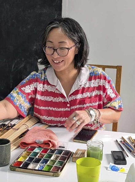 Una dona somrient amb ulleres està al costat de tres quadres abstractes emmarcats en una paret color crema.