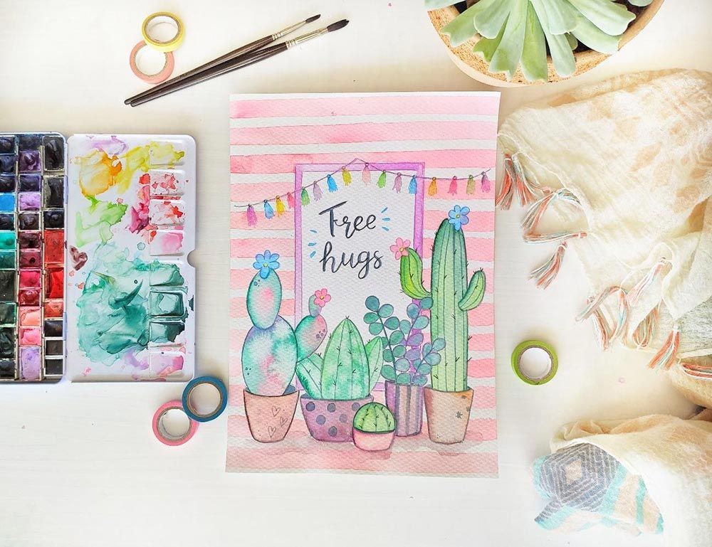 Materials d'art de cactus d'aquarel·la sobre un escriptori en tons pastel amb pinzells, cinta adhesiva i un quadern de dibuix que diu
