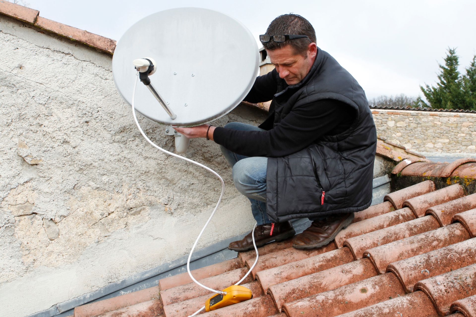 Un technicien, agenouillé sur un toit de tuiles, ajuste une antenne parabolique reliée à un indicateur de signal jaune.
