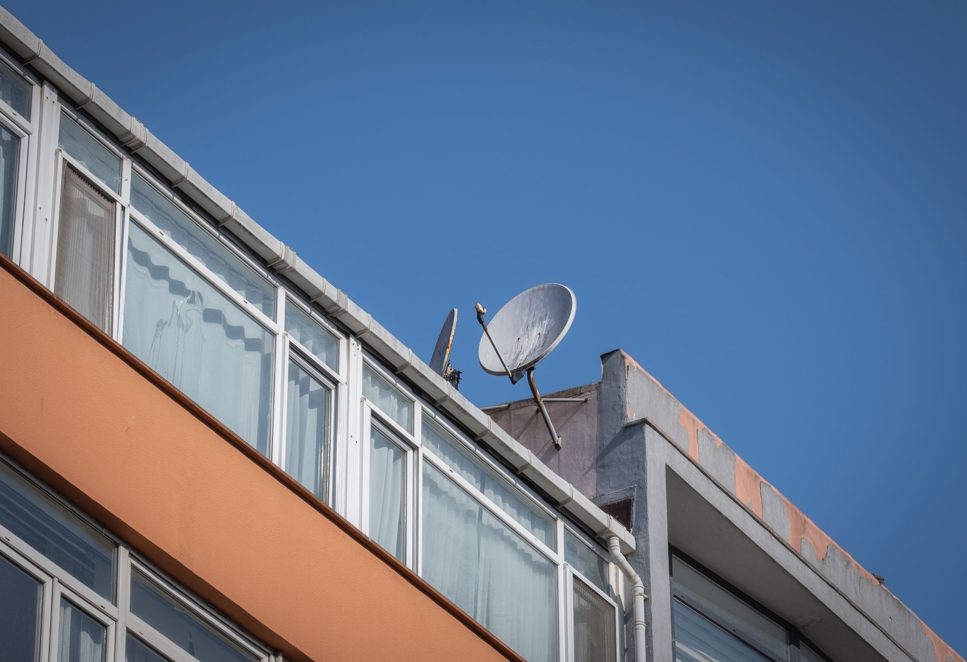 Une antenne parabolique est installée sur le toit plat d'un immeuble aux murs orange et aux balcons vitrés, sur fond de ciel bleu.