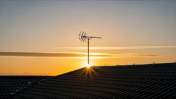 Une antenne de télévision se dresse sur un toit de tuiles sombres, se détachant en silhouette sur un lever de soleil doré et lumineux.
