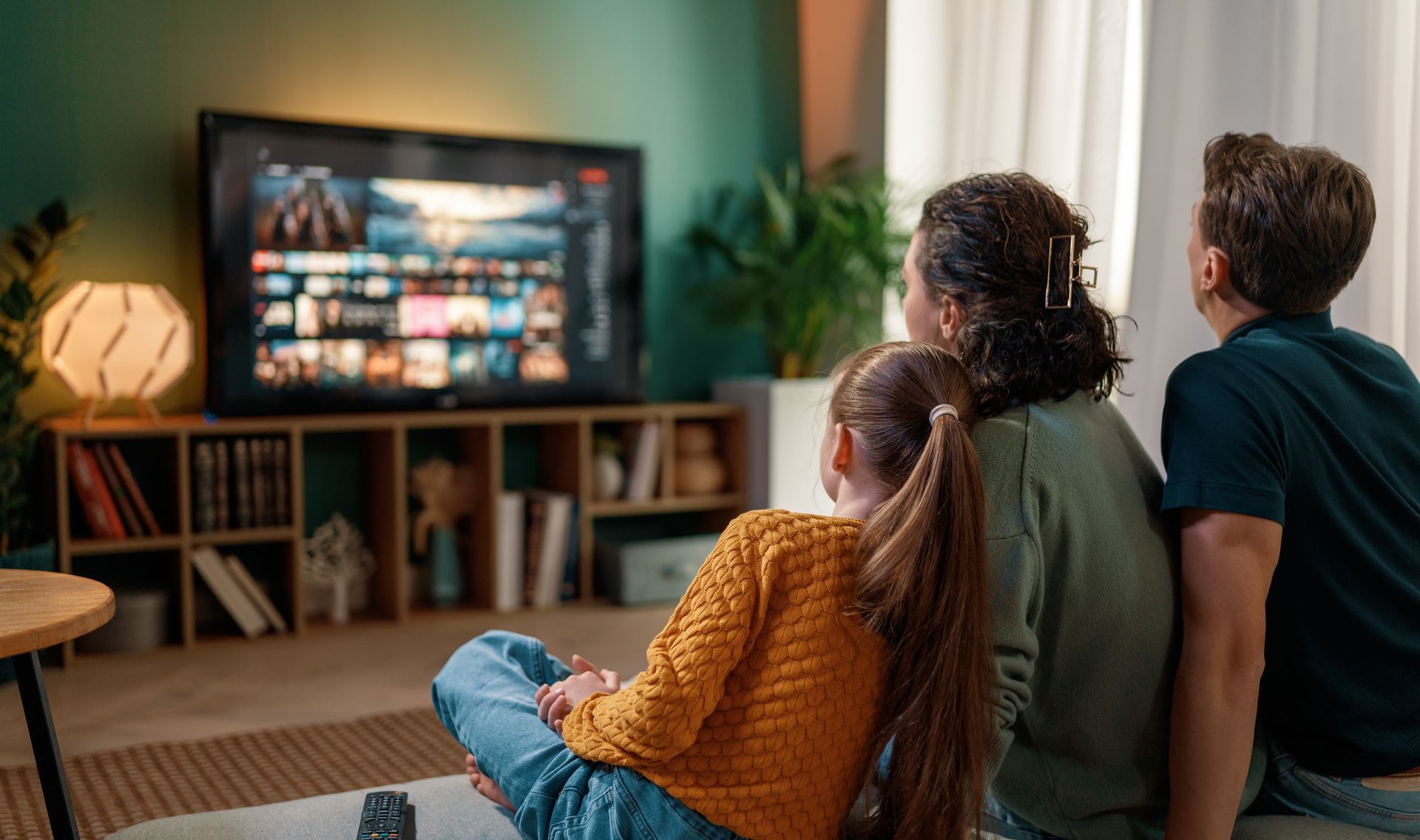 Trois personnes assises sur un canapé regardent un écran de télévision affichant un menu de films dans un salon confortable et faiblement éclairé.