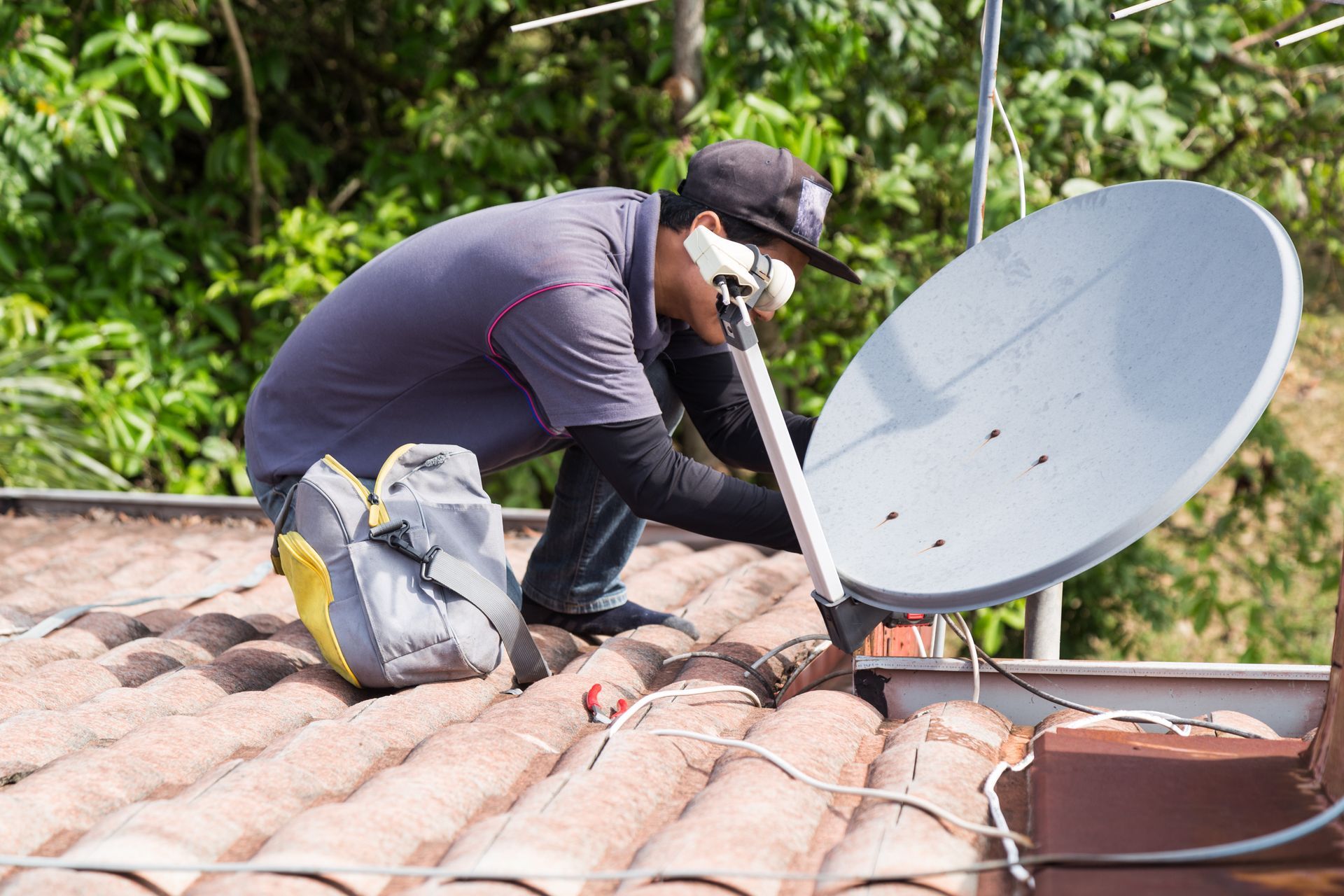 Un technicien en chemise grise, agenouillé sur un toit de tuiles, installe ou ajuste une antenne parabolique grise.