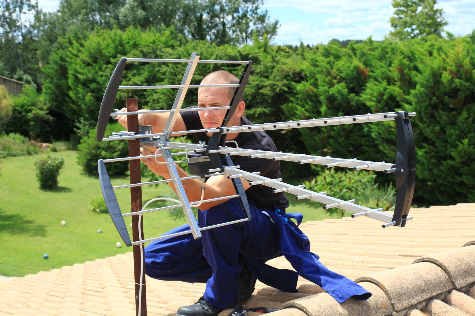 Un technicien en tenue de travail est accroupi sur un toit de tuiles pour installer ou régler une antenne de télévision métallique.