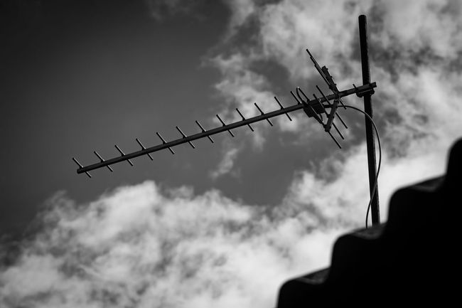 Une antenne de télévision fixée sur un mât, se détachant sur un ciel nuageux, photographiée en noir et blanc.