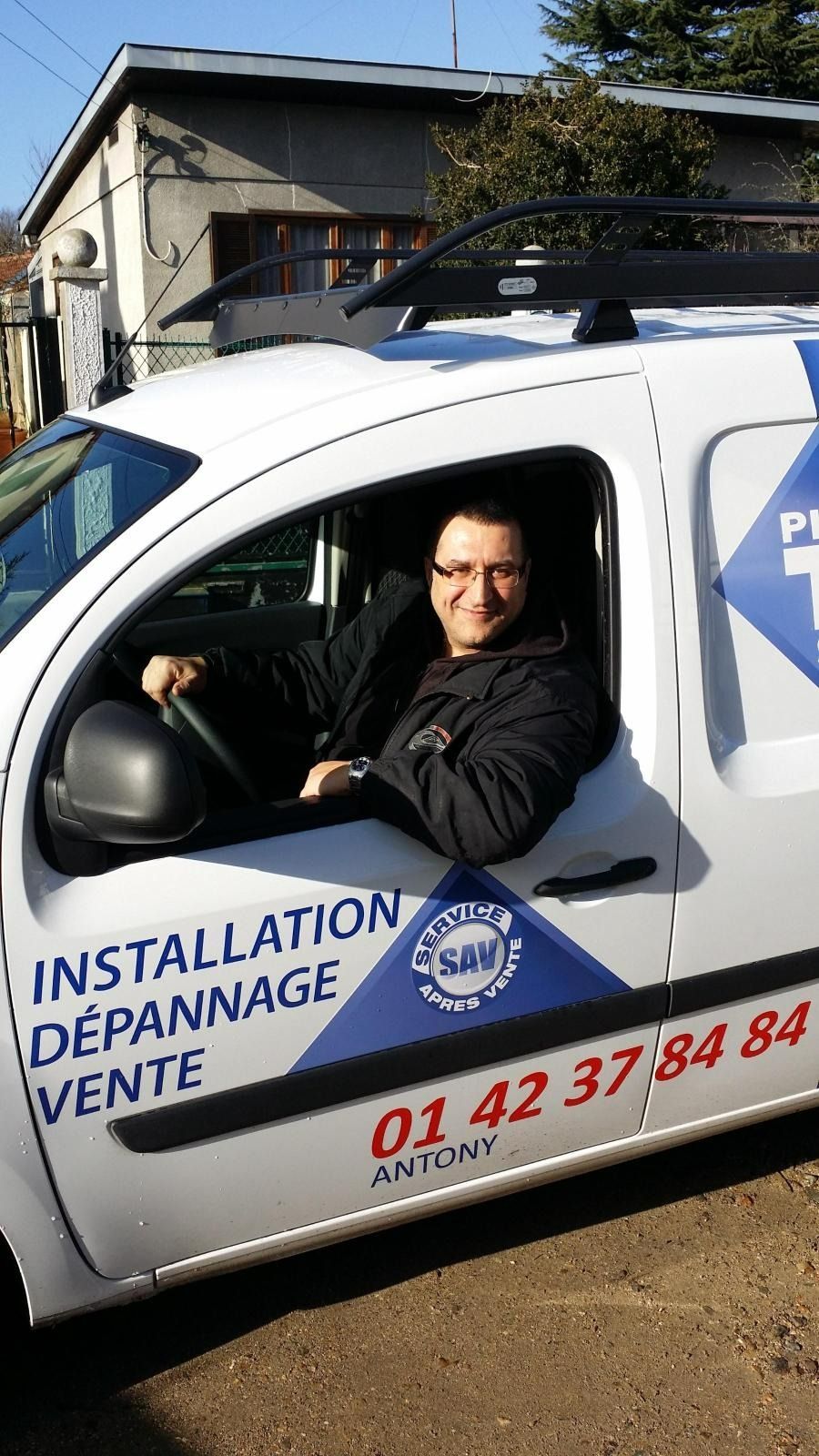 Une personne sourit depuis le siège conducteur d'une camionnette de service blanche avec des inscriptions en français et un numéro de téléphone sur le côté.