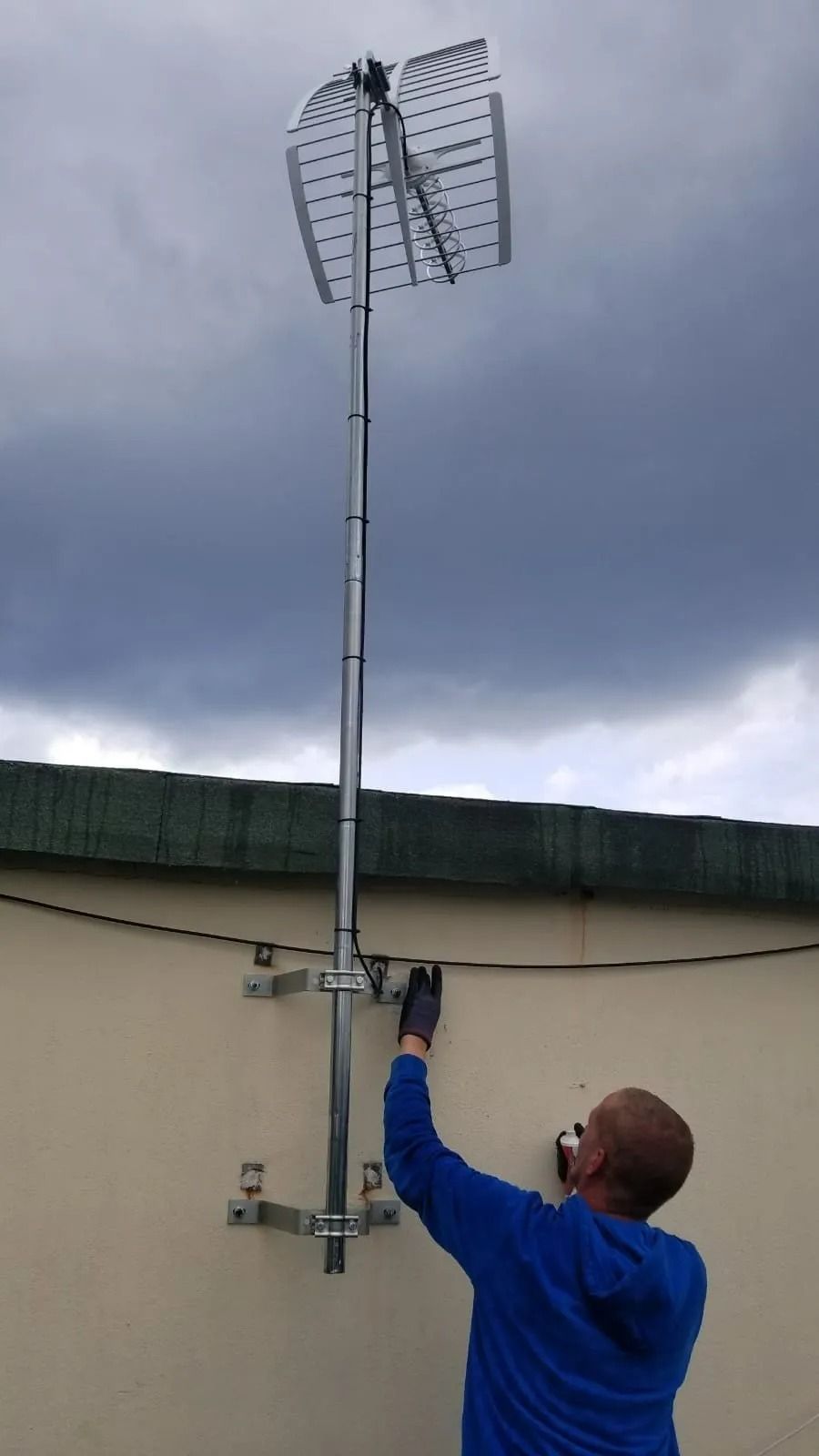 Une personne portant un sweat à capuche bleu installe une antenne de télévision sur un toit, sur un poteau métallique, sous un ciel nuageux.