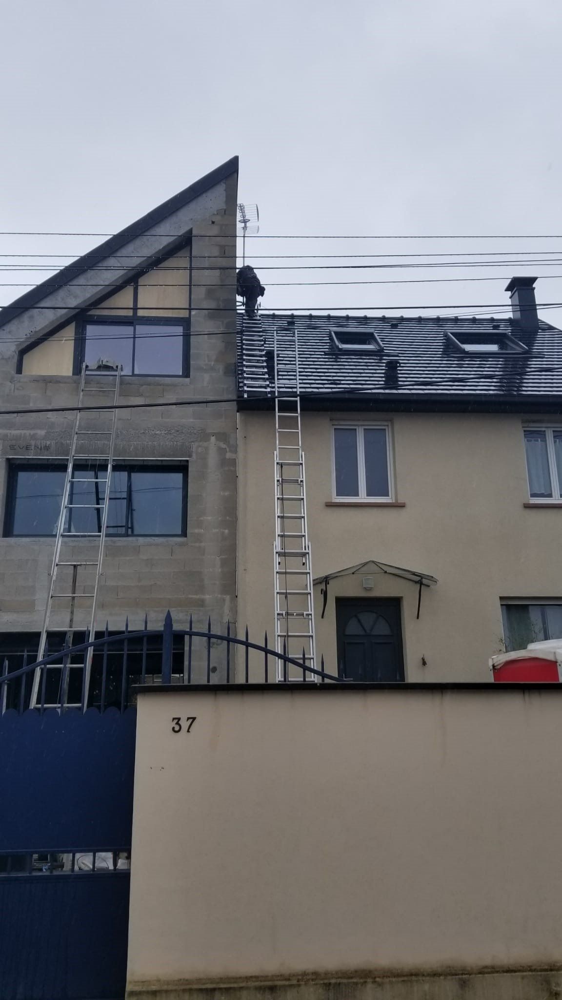 Deux maisons, dont une en construction, avec des échelles appuyées contre leurs murs, visibles derrière un muret et un portail bleu.