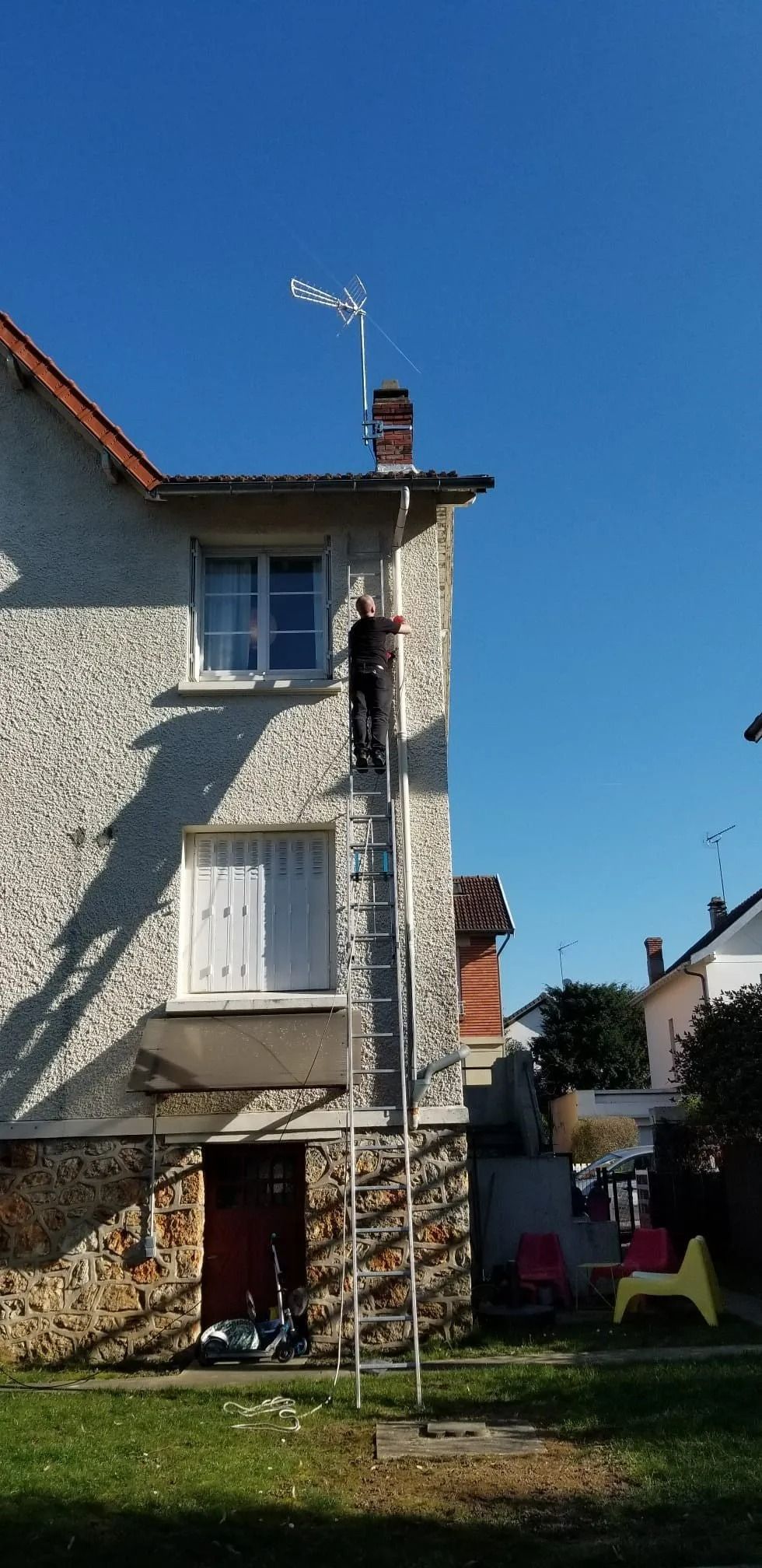 Par une journée ensoleillée, une personne, perchée sur une haute échelle, tend le bras vers le toit d'une maison en stuc.