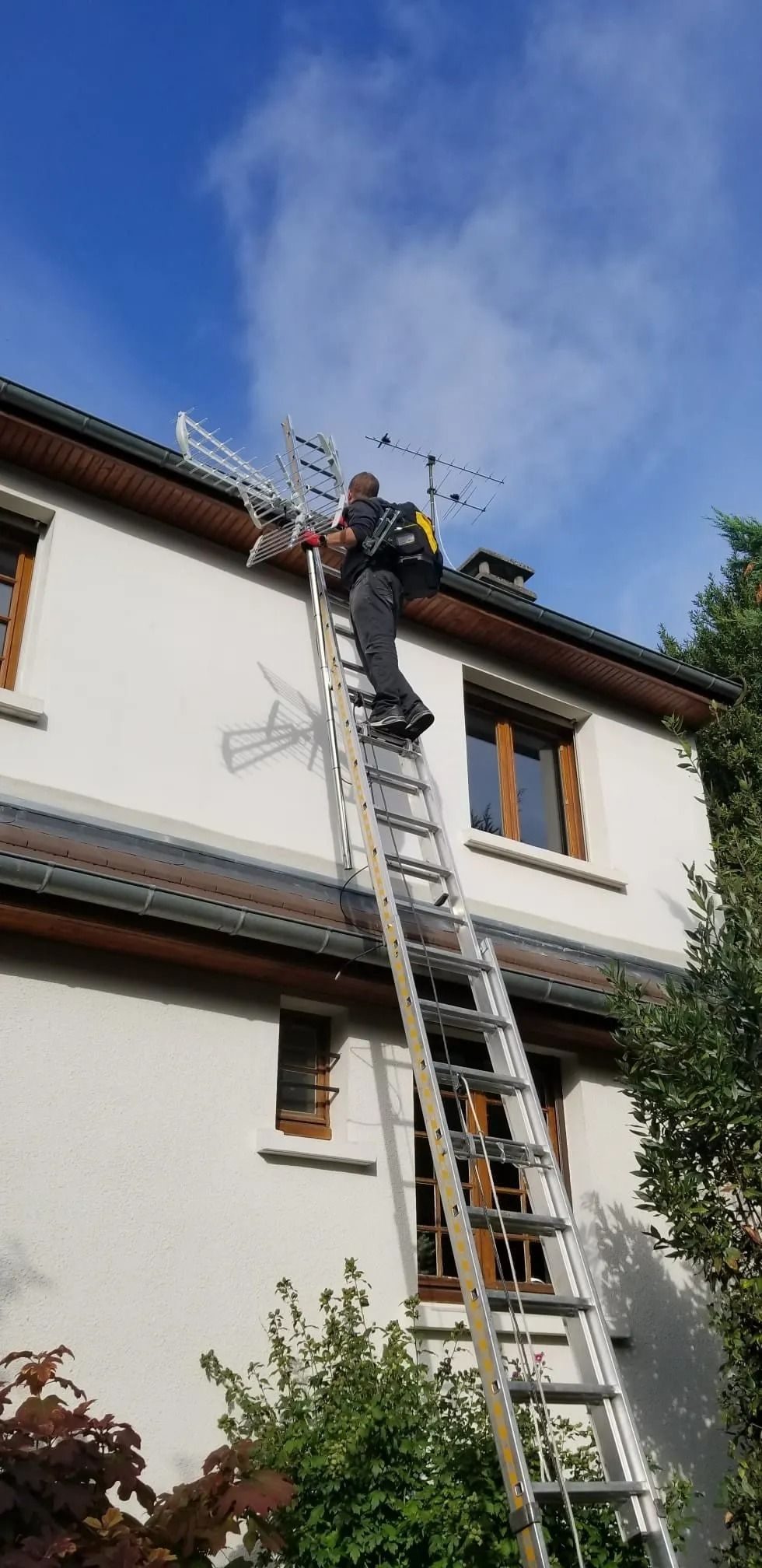 Une personne portant un sac à dos grimpe à une grande échelle métallique appuyée contre le mur blanc d'une maison à deux étages.