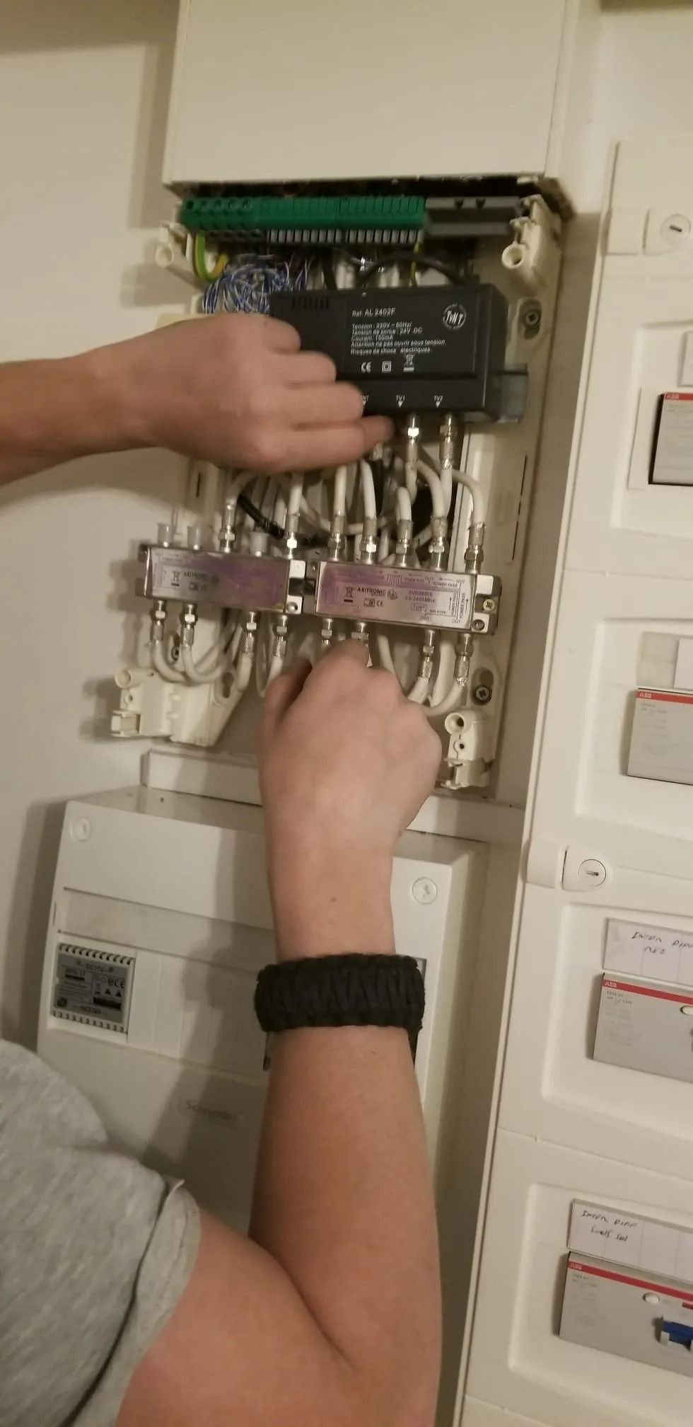 Les mains d'une personne ajustent des câbles blancs à l'intérieur d'un tableau de distribution électrique mural ouvert.