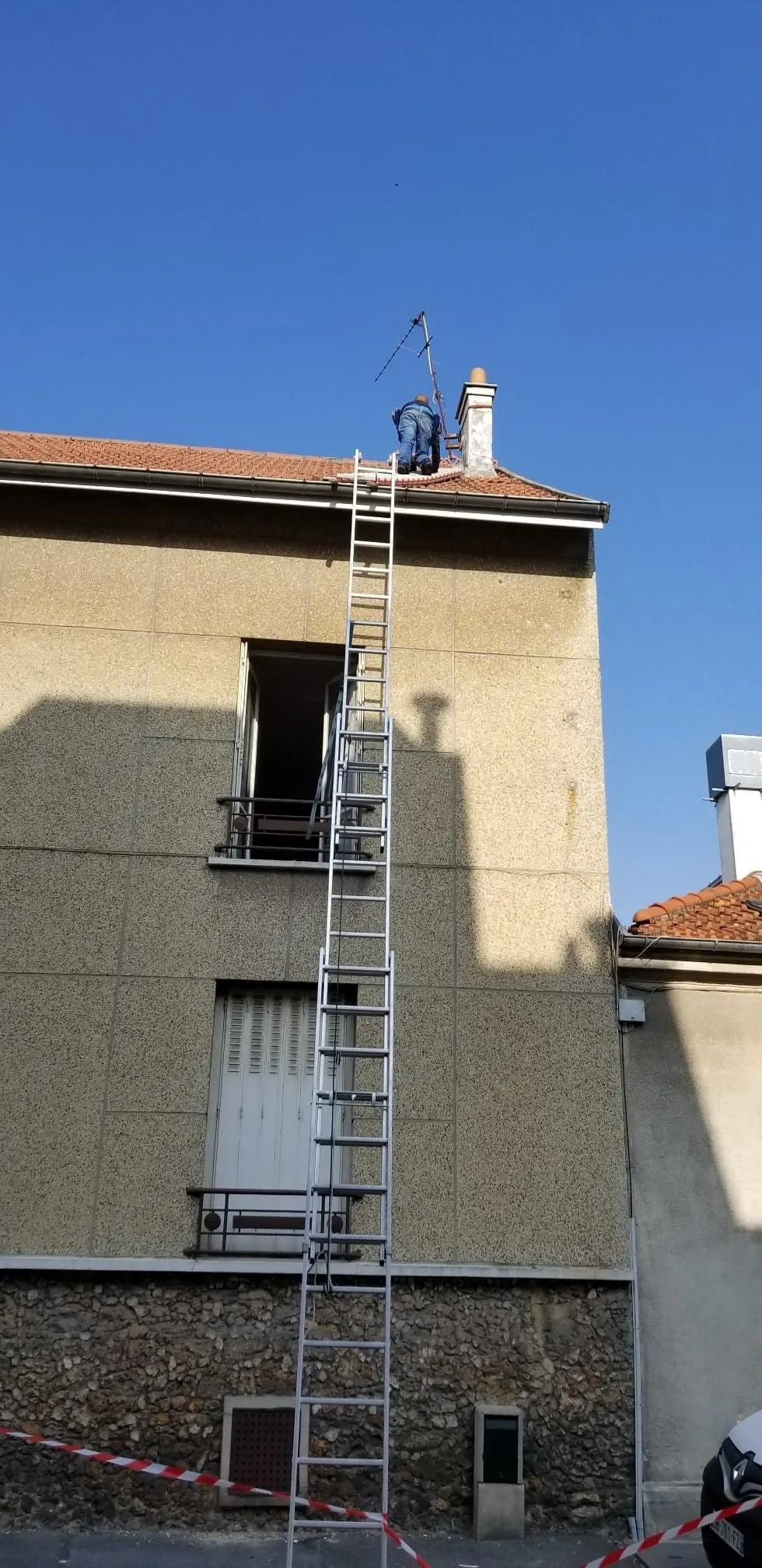 Sous un ciel bleu clair, une personne, perchée sur une haute échelle, tend le bras vers le bord du toit d'un bâtiment beige.