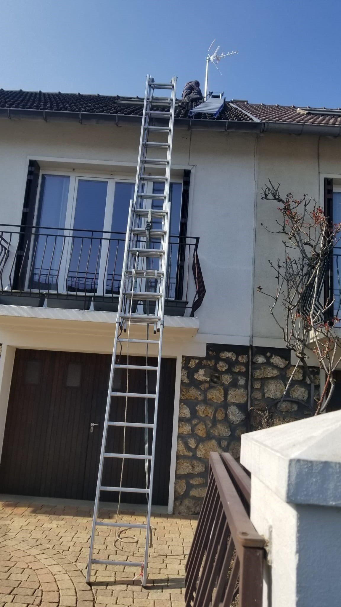 Une grande échelle métallique est appuyée contre le mur extérieur d'une maison blanche, tendant vers le toit et l'antenne de la cheminée.