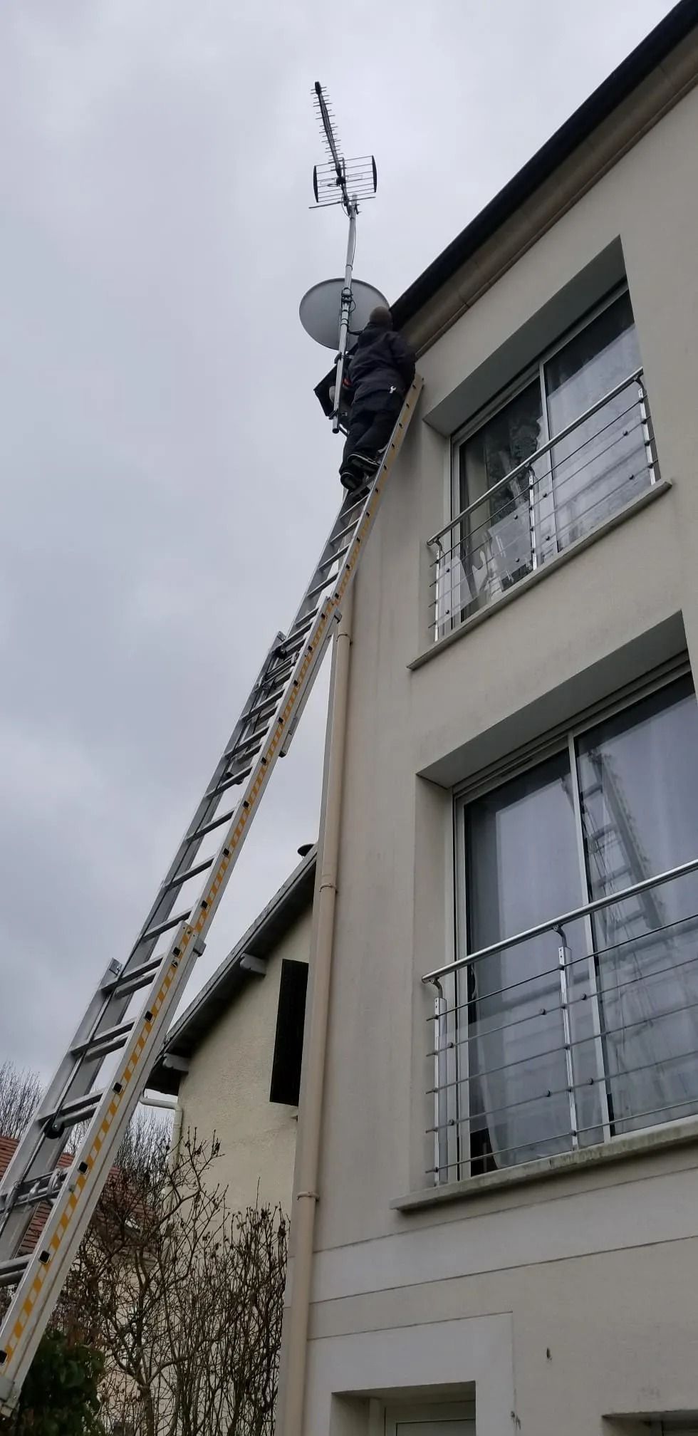 Un technicien, juché sur une échelle, ajuste une antenne de télévision et une parabole satellite fixées sur le côté d'un immeuble de plusieurs étages.