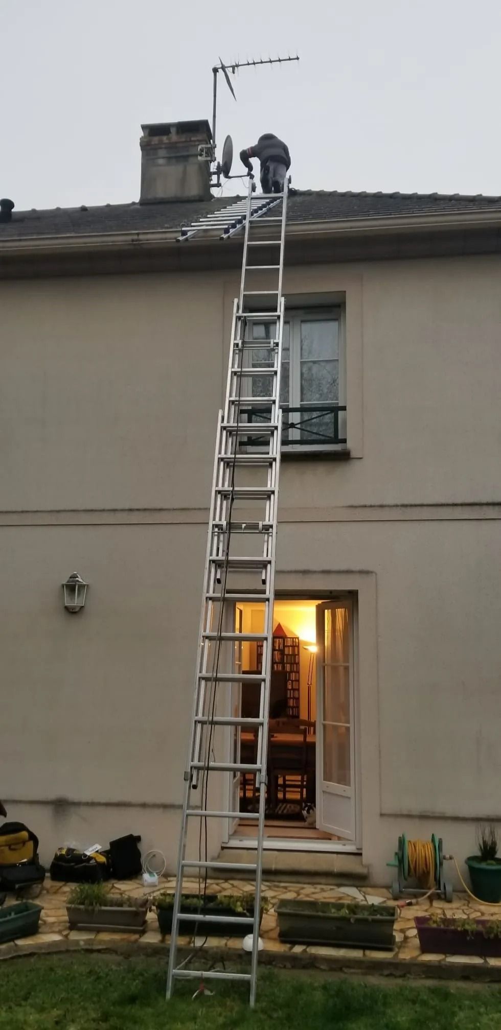 Une personne se tient sur une longue échelle appuyée contre le mur d'une maison à deux étages pour travailler sur le toit, près d'une cheminée.