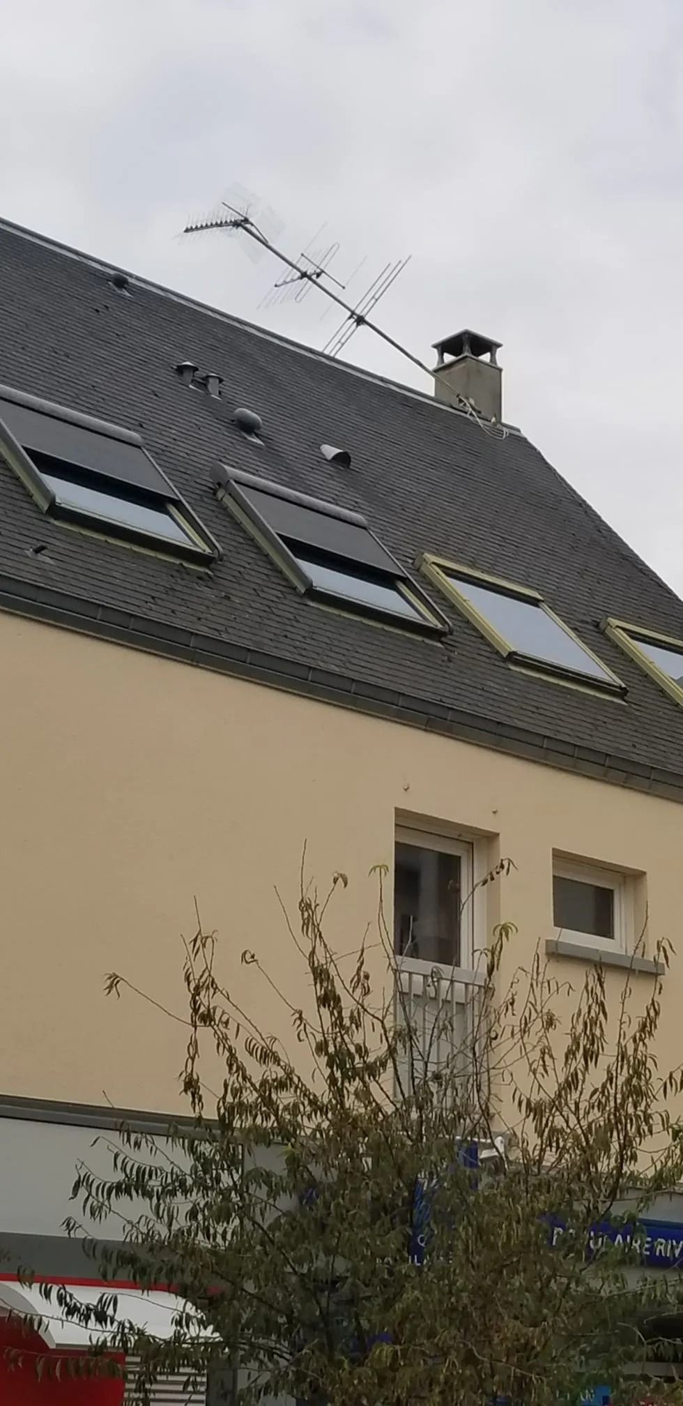 Une maison claire avec trois puits de lumière dans un toit gris foncé, une cheminée en briques et une antenne de télévision sur fond de ciel nuageux.