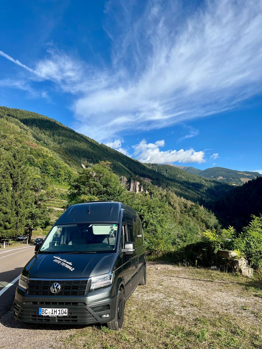 Ein schwarzer Lieferwagen ist am Rand einer unbefestigten Straße vor einem Berg geparkt.