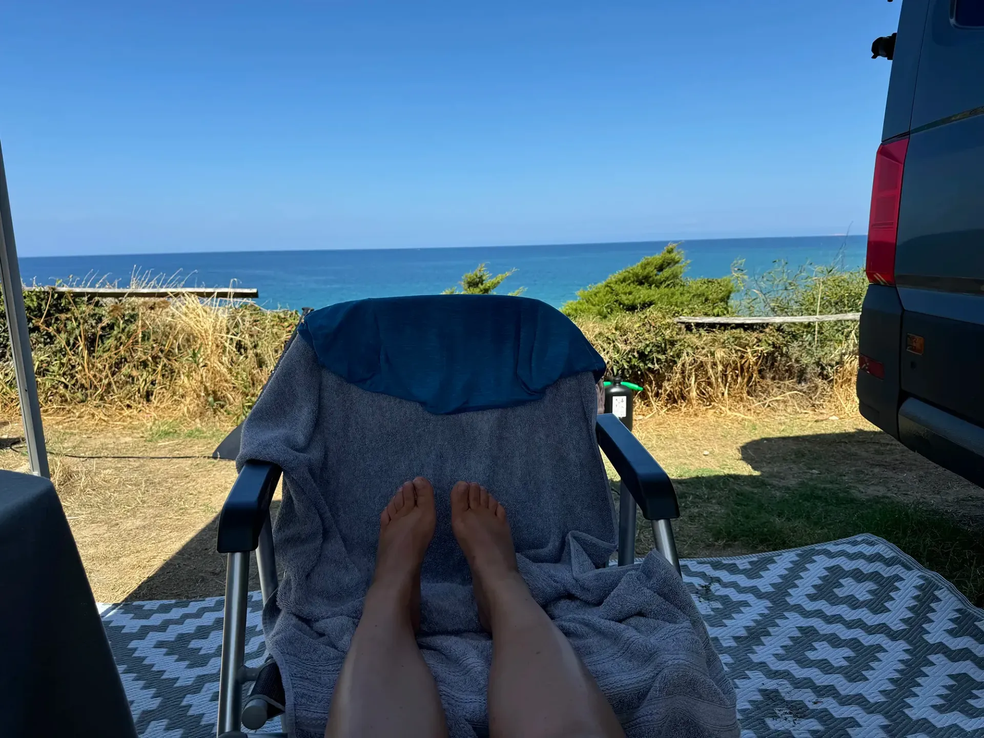 Eine Person sitzt auf einem Stuhl und hat die Füße vor dem Meer hochgelegt.