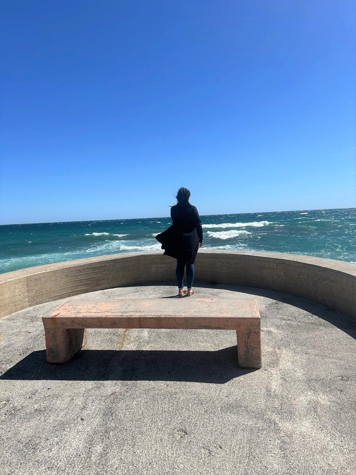 Eine Frau steht auf einer Bank und blickt auf das Meer.