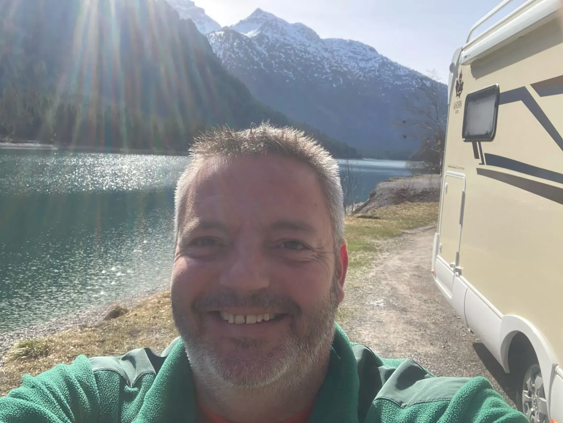 Ein Mann mit Bart lächelt vor einem See und Bergen.