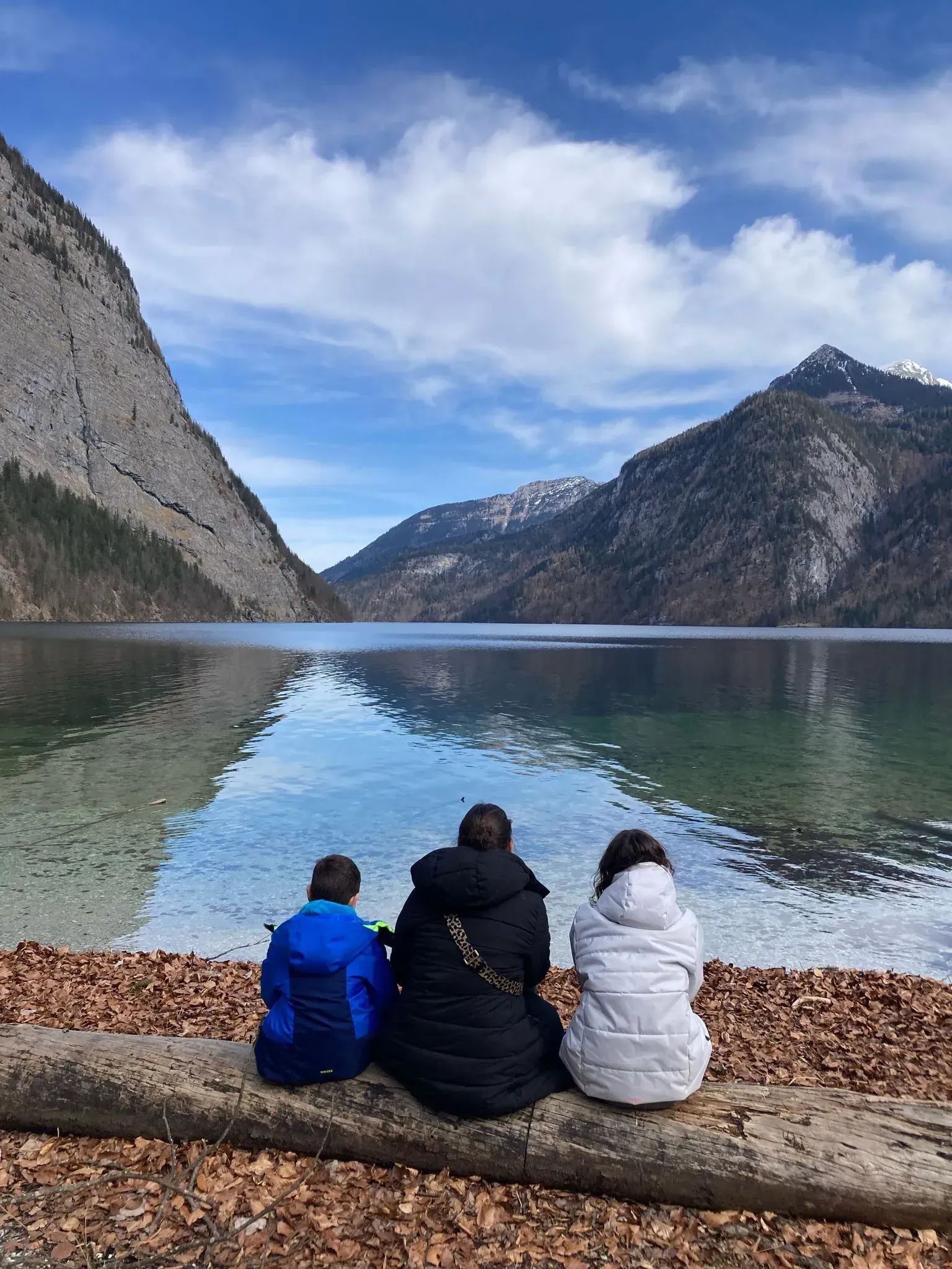 Drei Personen sitzen auf einem Baumstamm und blicken auf einen von Bergen umgebenen See.