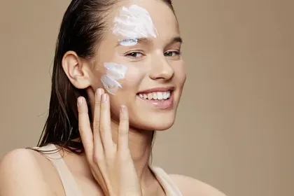 Dona aplicant-se crema blanca a la cara, somrient sobre un fons neutre.