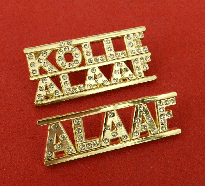 Goldfarbene Broschen mit klaren Strasssteinen, die „KOLLE ALAAF“ und „ALAAF“ auf rotem Hintergrund buchstabieren.