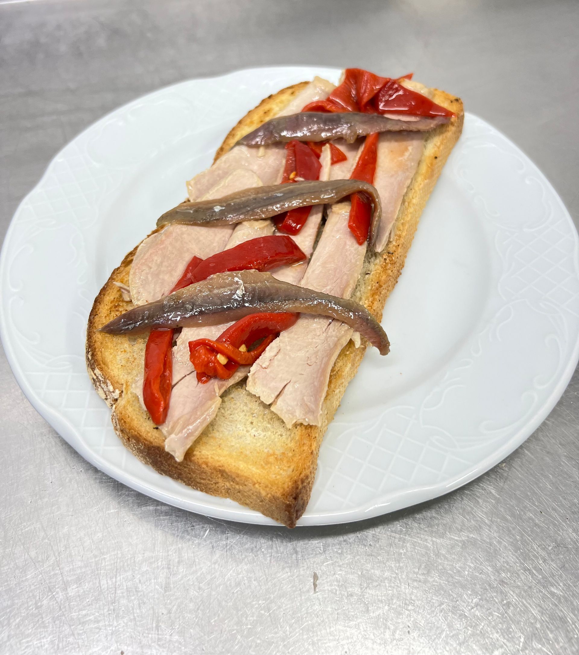 Pan tostado cubierto con atún, pimientos rojos y anchoas en un plato blanco.