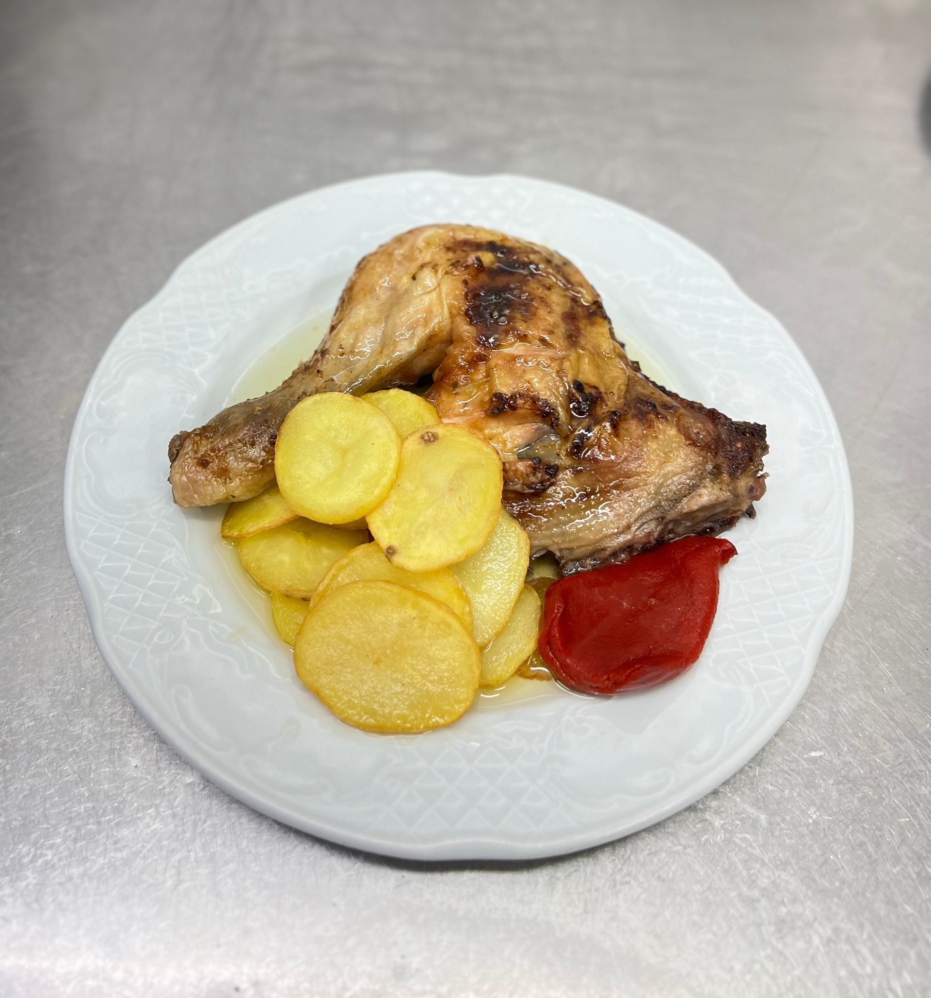 Muslo de pollo asado en un plato blanco con rodajas de patata y un pimiento rojo.