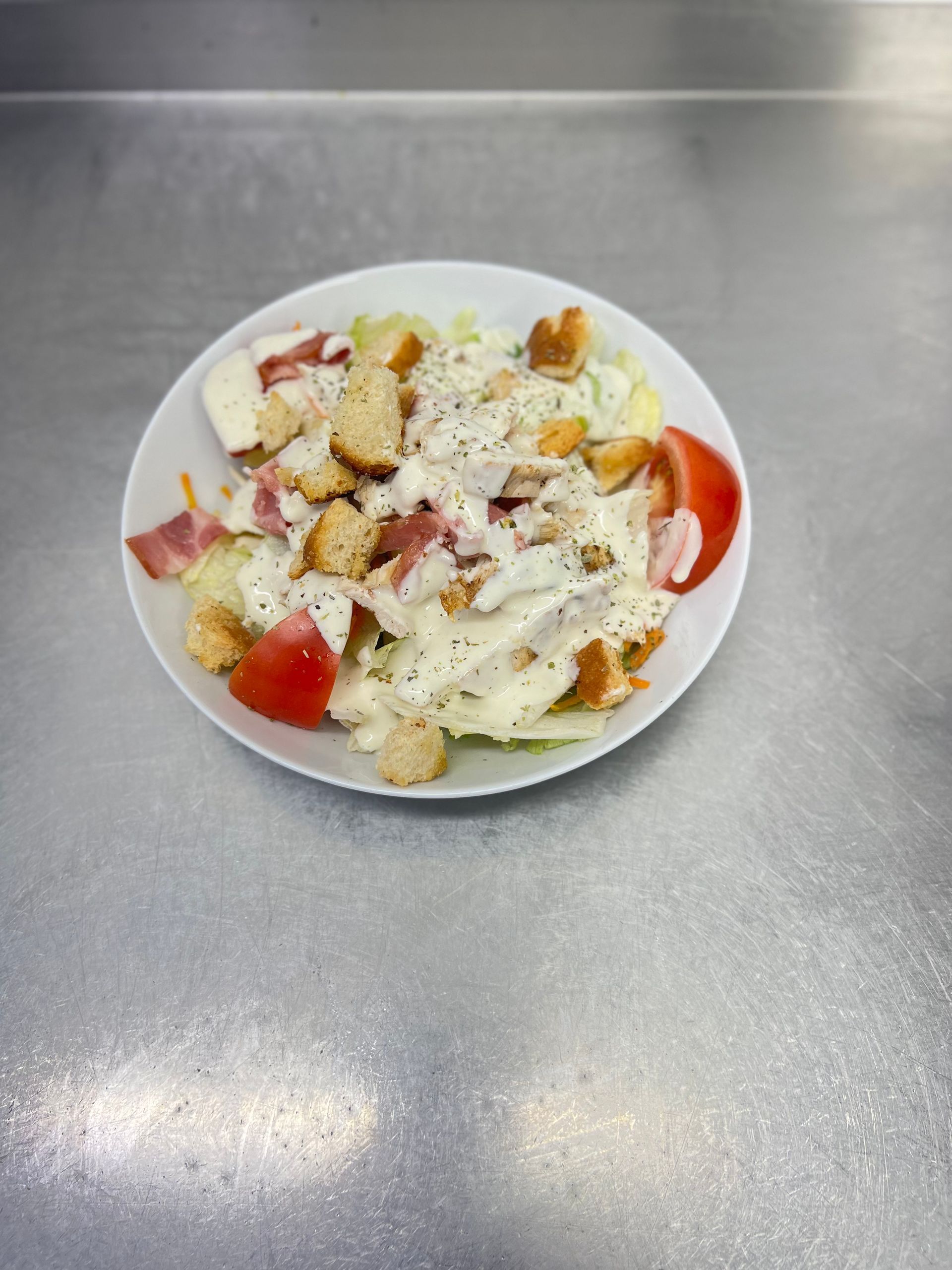 Ensalada César con crutones, tocino y tomate en un recipiente blanco sobre una superficie plateada.