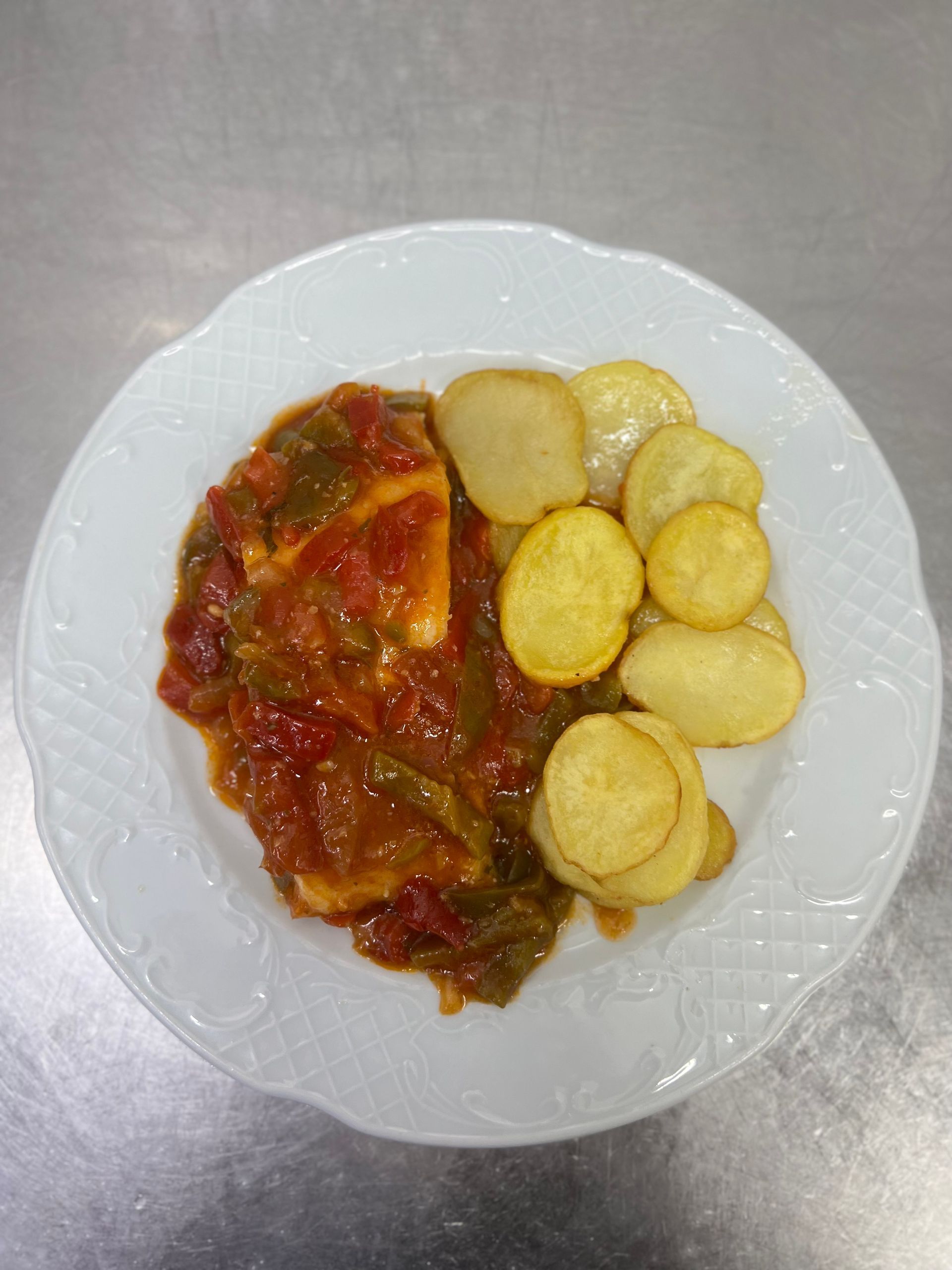 Filete de pescado en salsa roja con pimientos verdes, servido con rodajas de patatas fritas en un plato blanco.