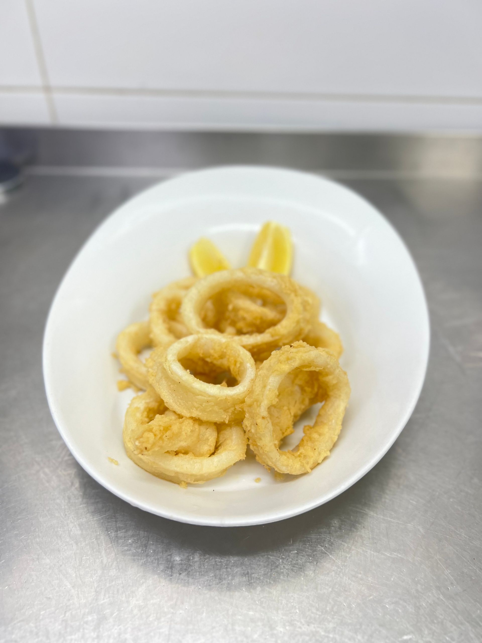 Anillos de calamares fritos en un plato blanco con rodajas de limón, apoyados sobre una superficie de acero inoxidable.