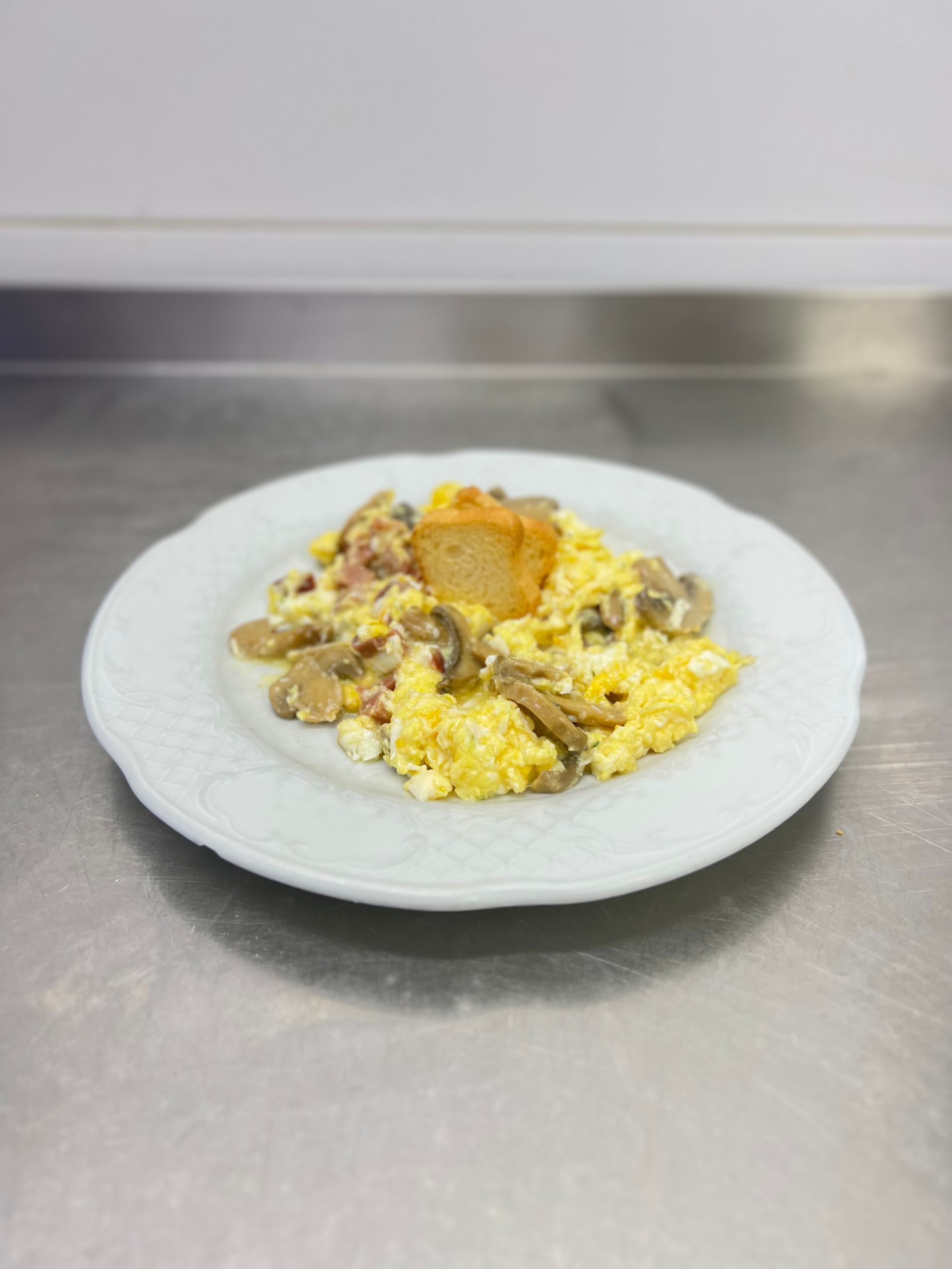 Huevos revueltos con champiñones, jamón y un crutón en un plato blanco, colocado sobre una superficie de acero inoxidable.