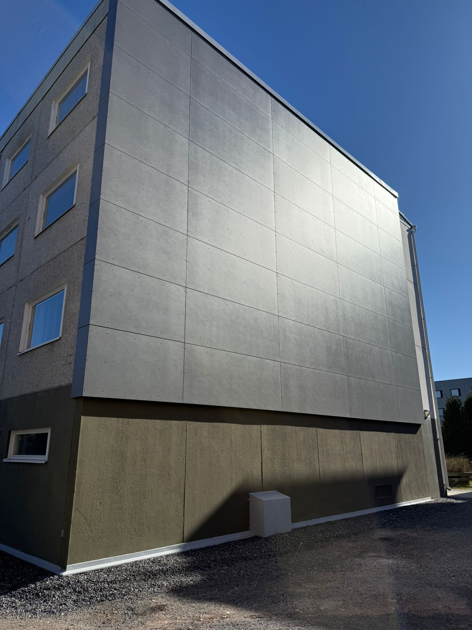 Moderni harmaa rakennuksen ulkoasu, jossa on tasainen seinä, pienet ikkunat ja sorapinta sinisen taivaan alla