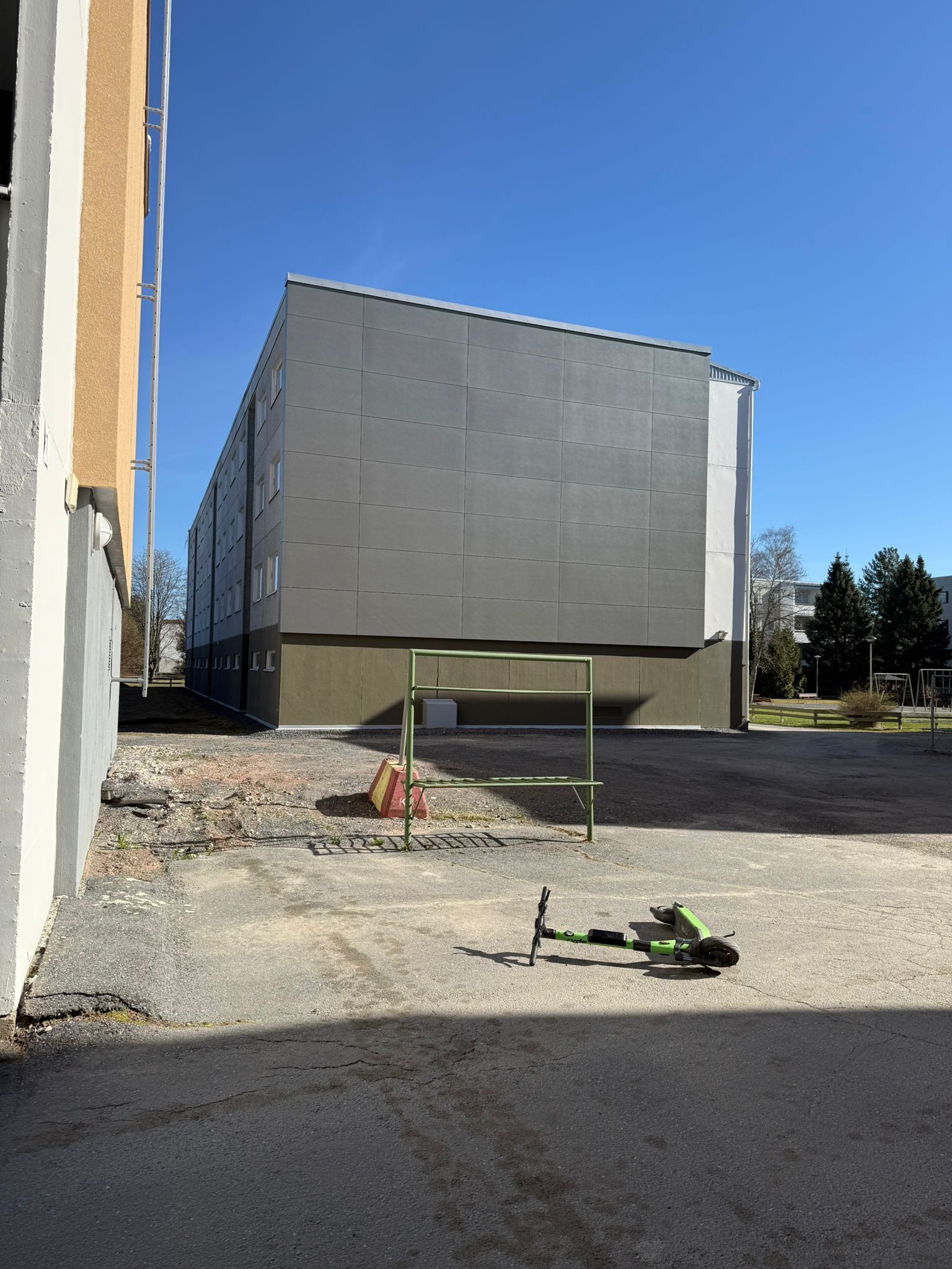 Harmaa rakennus soratolla toisen rakennuksen vieressä, etualalla pieni vihreä skootteri.