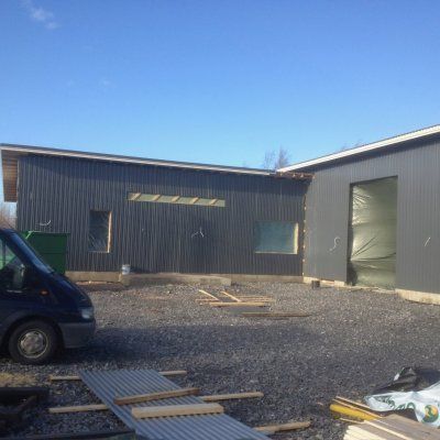 Sorainen piha tummanharmaan teollisuusrakennusten välissä kirkkaan sinisen taivaan alla, vasemmalla pakettiauto ja hajallaan olevia rakennusmateriaaleja.